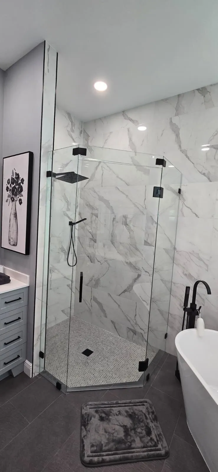 Frameless glass shower with black hardware beside a freestanding tub